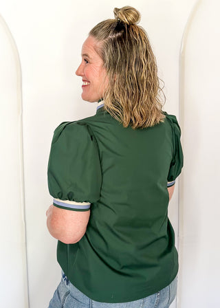 A woman with blonde hair in a bun is shown from the back wearing a dark green puff-sleeve blouse with striped cuffs, paired with light blue jeans, set against a white arched wall.