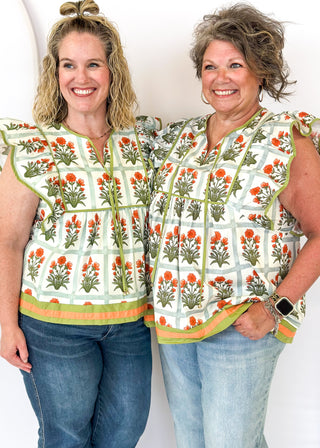 Flutter sleeve top with off white background and light green grid pattern-large orange florals with green stems/leaves in each grid. Olive green ribbon trim on sleeve and collar.  Olive green ribbon tie neck.  Olive green and orange trim on bottom band. 