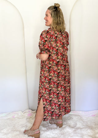A woman wearing a knee-length floral dress with short sleeves and a side slit, standing in a studio setup with white arched backdrops and a white faux-fur rug.