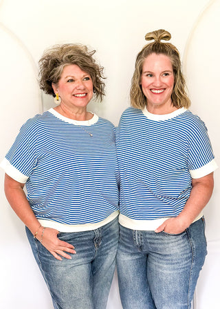 Two women wearing matching blue and white striped shirts and jeans standing against a white background.
