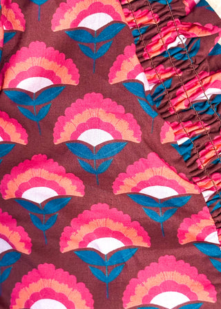 Close-up of a patterned fabric with a brown base and repeating pink, orange, and teal scalloped leaf motifs, plus a gathered ruffled section on the right.