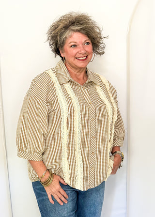 A smiling woman with short, curly gray hair models a beige and brown gingham button-up shirt with vertical cream lace stripes, paired with blue jeans, standing against a white arched studio backdrop.