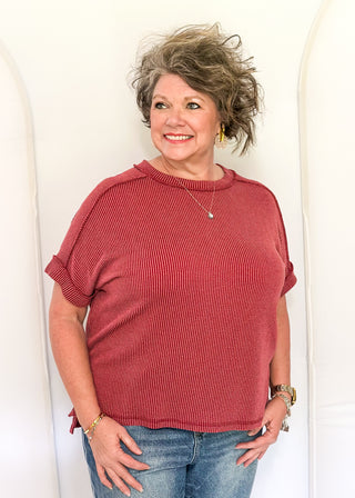 Woman wearing a red sweater and blue jeans standing against a white wall.