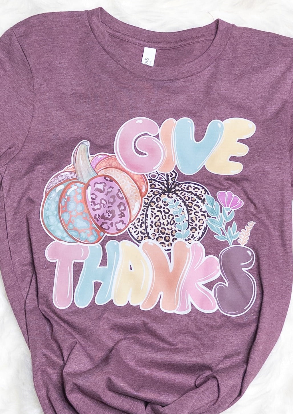 Deep mauve color tee shirt with bubble lettering that says "give thanks" in pale fall colors and 2 pumpkins. 1 pumpkin is animal print, the other is light fall color animal print.