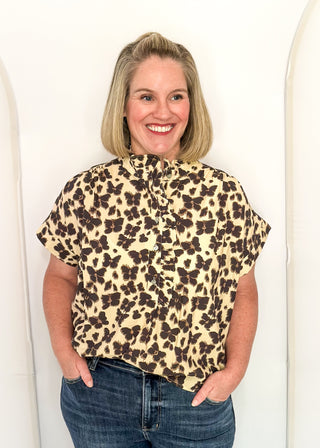A smiling woman with blonde hair wearing a short-sleeve leopard print blouse and blue jeans, standing against a white arched wall.