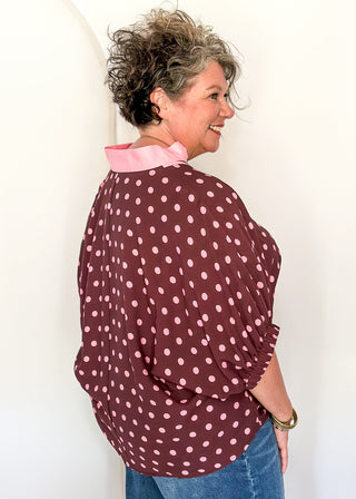 A woman wearing a burgundy and pink polka-dot blouse with a light pink collar, paired with blue jeans, standing against a white arched backdrop.