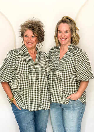 Two women standing side-by-side wearing matching green and white gingham short-sleeve button-up shirts and blue jeans, posing against a white arched backdrop.
