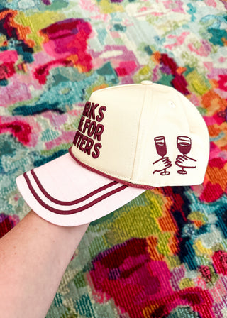 A cream-colored baseball cap with burgundy lettering that reads 'WORKS FOR WANTS' and a graphic of two clinking wine glasses on the side, held by a hand against a colorful patterned rug.