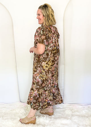 A woman wearing a brown floral midi dress with short puff sleeves and a ruffled hem, standing in front of a white arched backdrop on a white faux fur rug.