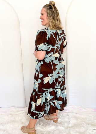 woman standing profile in a brown, soft blue and off white floral midi dress.