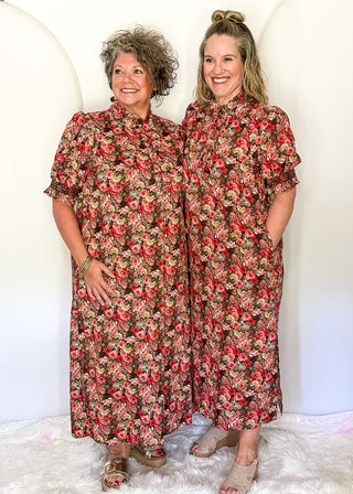Two women standing side by side wearing matching short-sleeve floral maxi dresses with pockets, featuring a red, pink, and green floral pattern on a dark background, posed against a white studio backdrop with a white faux fur rug.
