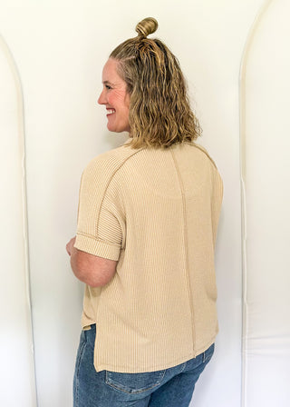 Person wearing a beige ribbed sweater and blue jeans standing against a white wall.