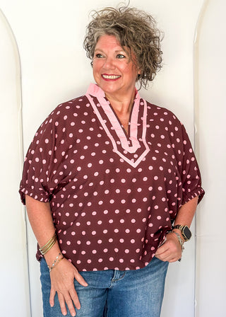A smiling woman with curly gray hair wearing a burgundy short-sleeve top with pink and white polka dots and a pink V-neck trim, paired with blue jeans, standing against a white arched wall.