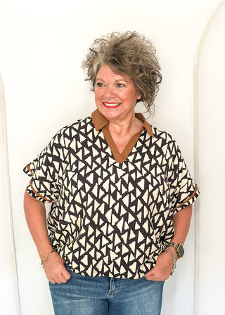A woman with short, curly gray hair wearing a short-sleeve patterned blouse with blue jeans, standing against a white arched wall.