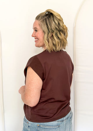 A woman with blonde hair tied back is shown from the back wearing a short-sleeve brown t-shirt and light blue jeans, standing against a white studio backdrop with arched panels.
