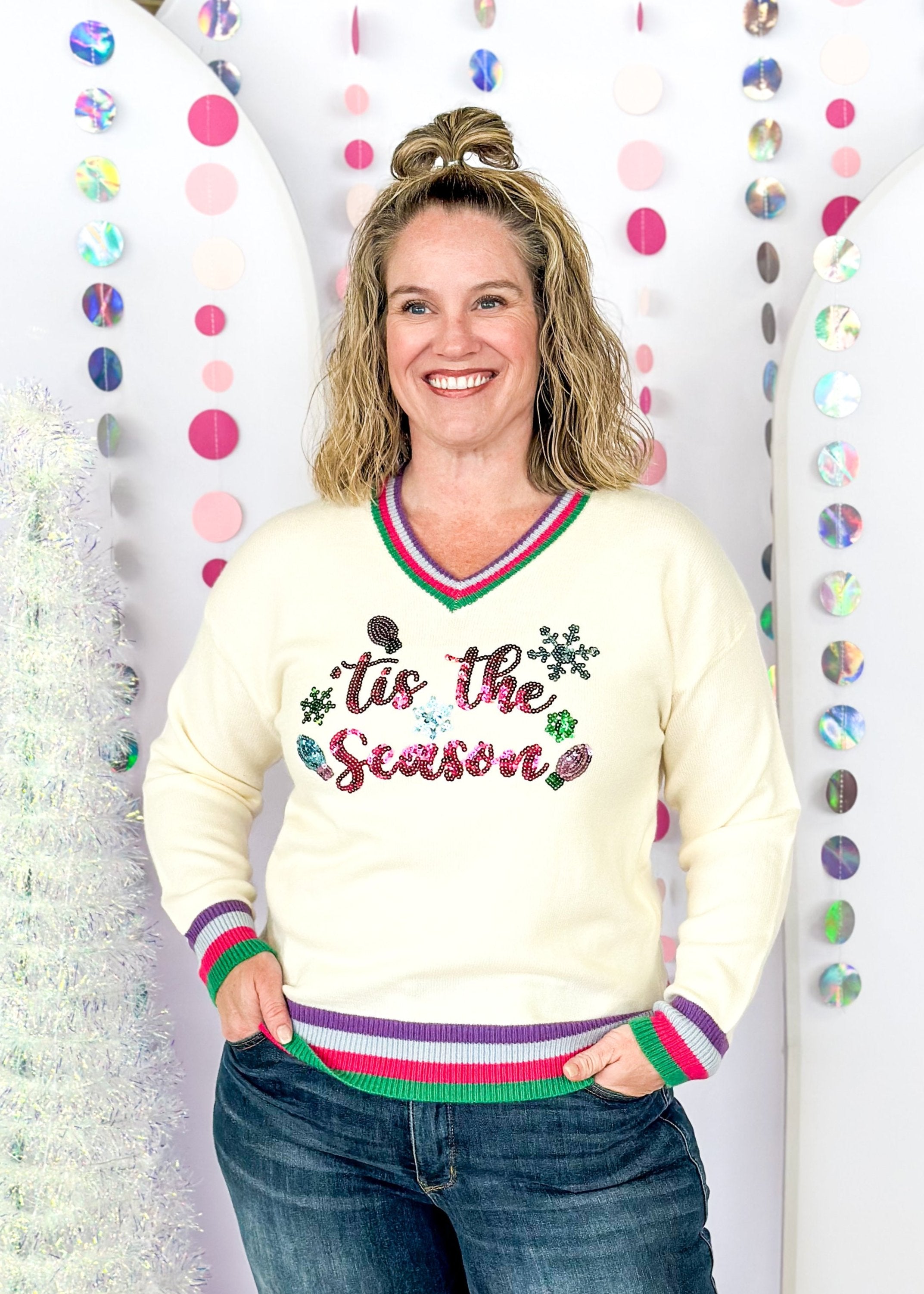 cream long sleeve sweater with v-neck, cuffs and bottom banding all striped in purple, pink, green and light blue, tis the season spelled out across the front in sequins