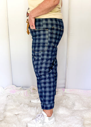 A person shown from the waist down wearing blue and gray buffalo plaid cropped pants, a pale yellow top, and white sneakers, standing on a white faux fur rug against a white studio backdrop.