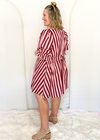 woman standing profile, wearing pink and wine striped dress with pockets. 