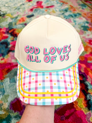A cream-colored snapback cap held by a hand, featuring the text 'GOD LOVES ALL OF US' in pink and teal bubble letters on the front. The brim has a pastel pink, blue, and white gingham pattern decorated with small yellow stars, and is outlined with yellow piping.