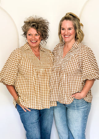 Two women standing side-by-side wearing matching tan and white gingham short-sleeve button-up shirts and blue jeans, posing in front of a white studio backdrop with arches.
