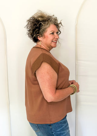 A woman with short, messy gray-blonde hair is shown from the side, wearing a sleeveless brown top and blue jeans, standing against a white studio backdrop with arch-shaped supports.
