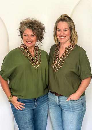 Two women standing side-by-side wearing matching olive green short-sleeve tops with a leopard-print V-neck trim, paired with blue jeans.