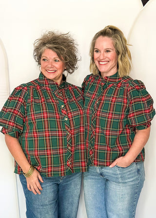 Two women standing side by side wearing matching green and red plaid puff-sleeve button-up shirts with blue jeans, posing in front of a white arched backdrop.