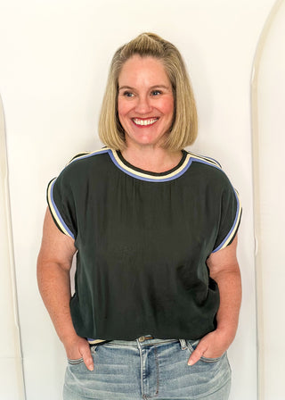 A smiling blonde woman wearing a dark olive green short-sleeve top with light blue and white striped trim on the collar and sleeves, paired with light-wash blue jeans, standing against a white arched wall.