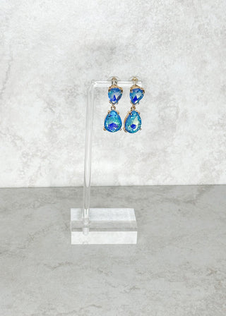 Blue teardrop earrings on a clear stand against a light gray marble background