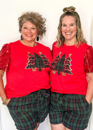 Two women wearing matching red shirts with Christmas tree designs and plaid shorts.