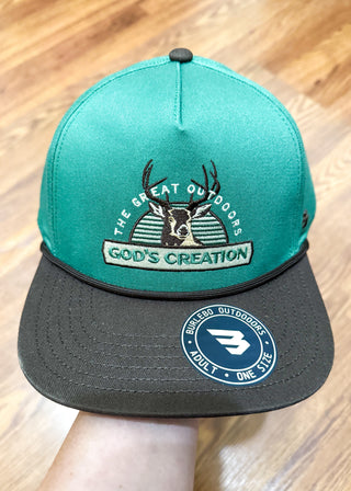 A teal and black snapback cap held by a hand over a wooden floor background. The front of the cap features an embroidered graphic of a deer head with antlers and the text 'THE GREAT OUTDOORS' and 'GOD'S CREATION'.