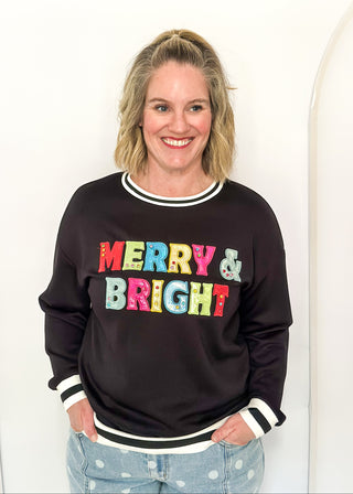 A smiling blonde woman wearing a black crewneck sweatshirt with 'MERRY & BRIGHT' printed across the chest in colorful, patterned letters, paired with light blue polka dot jeans against a white studio background.