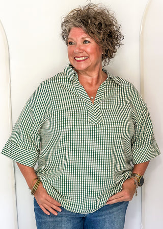 A smiling woman with short curly gray-brown hair stands against a white arched backdrop, wearing a green and white gingham short-sleeve button-up shirt with blue jeans.