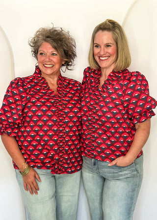 Two women standing side-by-side wearing matching red and black patterned short-sleeve button-up shirts and light-wash blue jeans, posing against a white arched studio backdrop.
