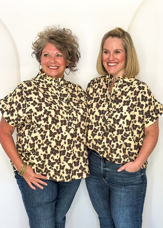 Two women standing side by side wearing matching leopard-print short-sleeve collared shirts and blue jeans, posing against a white arched wall.