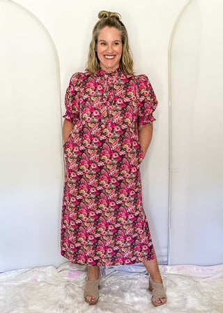 A woman stands in front of a white arched backdrop wearing a pink and red floral midi dress with puff sleeves and side pockets, paired with beige heeled sandals.