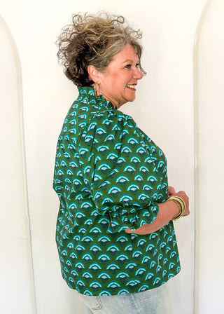 A woman with curly gray hair is shown from the side, wearing a green long-sleeve button-up shirt with a repeating light blue and white arch pattern, paired with light-wash jeans, standing against a white arched backdrop.