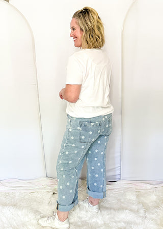A woman stands in profile wearing light blue denim jeans with white polka dots, a white t-shirt, and white sneakers, standing on a white faux fur rug in front of white arch backdrops.