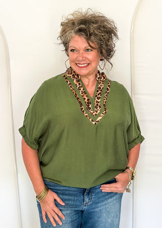A smiling woman with curly gray hair wearing an olive green short-sleeve top with a leopard-print V-neck collar, paired with blue jeans, standing against a white arched backdrop.