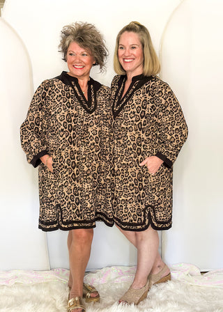 Two women standing side by side wearing matching leopard-print short robes with black trim, posed against a white studio backdrop with arch shapes and a white faux-fur rug.