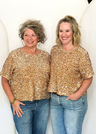 Two women standing side by side wearing matching short-sleeve sequined tops in a gold and beige pattern, paired with blue jeans, against a white arched backdrop.
