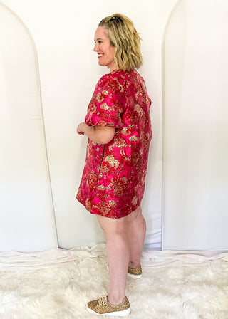 A woman stands in profile wearing a short pink dress with a gold floral pattern, paired with leopard-print slip-on shoes, set against a white arched backdrop on a white faux fur rug.