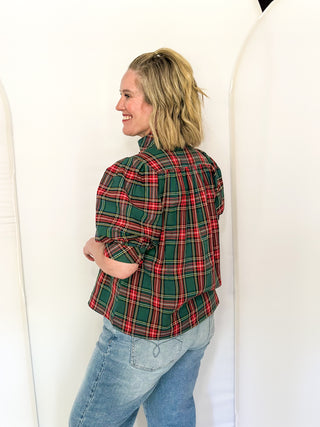 A woman with blonde hair tied back is shown from the back wearing a green and red plaid flannel shirt and light blue jeans, standing against a white studio backdrop.