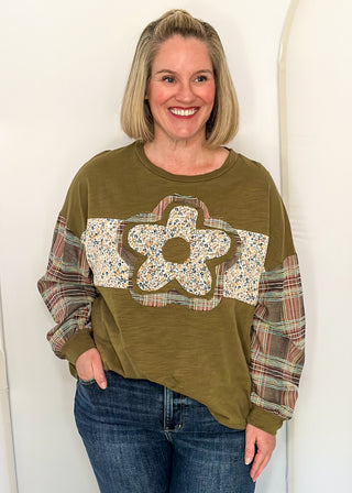 woman wearing an olive green  patchwork top with floral fabric pattern on the front and plaid long sleeves. 