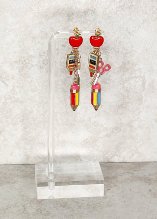 Dangle earrings with an apple, scissors, glue, and pencil displayed on a stand 