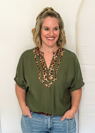A woman with blonde hair stands against a white studio backdrop wearing an olive green short-sleeve blouse with a leopard-print V-neckline detail and light blue jeans.