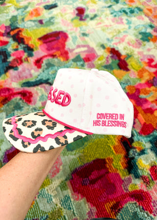 A hand holding a white baseball cap with pink polka dots, pink embroidered text reading 'COVERED IN HIS BLESSINGS,' and a leopard-print brim with a pink ruffled edge, set against a colorful patterned rug.