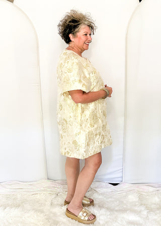 An older woman stands in profile wearing a short-sleeve cream or pale yellow lace-patterned dress with gold sandals, posing in front of a white arched backdrop.