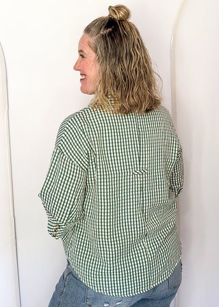 A woman with blonde hair in a bun is shown from the back, wearing a green and white gingham long-sleeve shirt and blue jeans, standing against a white wall with arched details.