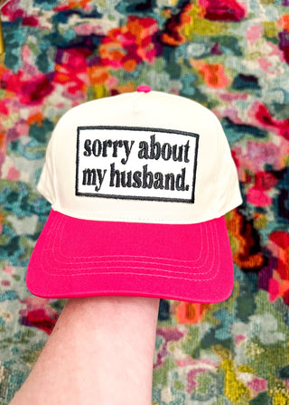 Cream snap back ball cap with a hot pink bill and embroidered patch “sorry about my husband,” stitched in black.